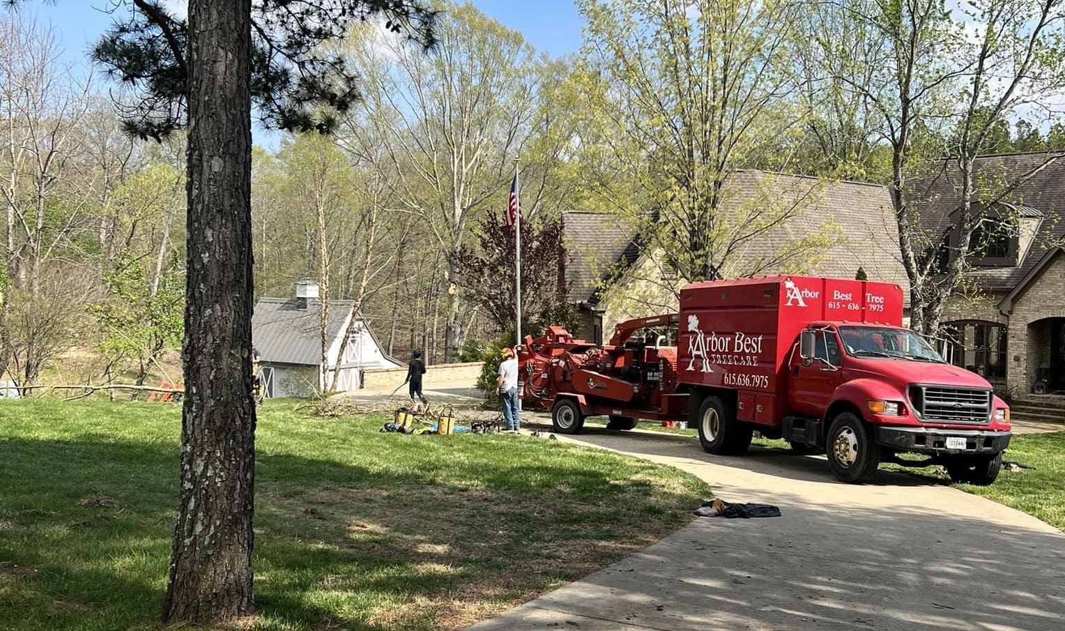 Tree Service A red Arbor Best tree service truck with a wood chipper is parked in the driveway of a house surrounded by trees. Arborists are seen handling equipment and working near the truck on a sunny day. Middle Tennesseer